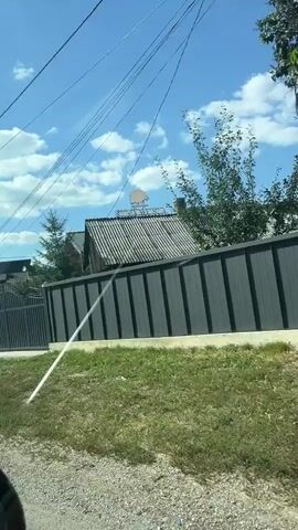 This is a first-person perspective video taken from a vehicle moving through a rural area on a sunny day. The camera pans across various scenes, including houses with fences, trees, power lines, and a street. There are signs visible, one of which reads "PENSION POPESCU". In one scene, two black cars are parked on a grassy area with a white line marking a sidewalk. Another scene shows a stop sign and a yield sign at an intersection. The overall impression is of a scenic drive through a residential village.