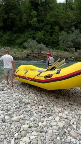 The media file depicts a group of people preparing to launch a yellow inflatable raft on a rocky riverbank. In the foreground, a man in a white t-shirt and shorts stands beside the raft, holding onto a rope attached to it. Another man, wearing a dark t-shirt and shorts, is positioned behind the raft, appearing to help push it. Inside the raft, a person wearing a red helmet is seated and holding a paddle.

The scene is set by a clear, turquoise river with a gentle current, bordered by lush green trees and vegetation on the opposite bank. The ground is covered with smooth, grey and white pebbles, indicating a natural, undeveloped riverside location. The sky appears overcast, suggesting a cloudy or potentially damp day, but the overall atmosphere is one of outdoor adventure and preparation. The activity is clearly the launch of a rafting trip, with the participants getting ready to enter the water. There are no visible text elements in the media file. The presence of the raft, paddles, and helmets strongly suggests a recreational activity like white-water rafting.