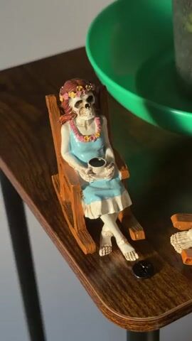 This is a close-up, high-angle shot of two small skeleton figurines sitting in rocking chairs on a wooden side table. The figurine on the left is a female skeleton wearing a blue dress with white trim and a floral lei around its neck and a flower crown on its head. It is holding a small cup of what appears to be coffee. The figurine on the right is a male skeleton wearing a white garment and is also holding a small cup. Both skeletons are positioned in rocking chairs with a coffee cup in hand, suggesting they are relaxing and possibly conversing. In the background, a green plant pot with dark soil and green stems is visible. The lighting suggests it is daytime, with sunlight casting shadows on the table and figurines. A small black button or coin is on the table between the two figurines.