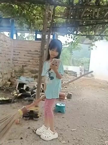 A young girl stands holding a broom. She is wearing a light blue t-shirt, pink leggings, and white clogs. She holds a pink cell phone in her left hand. She swings the broom from side to side as if pretending to fly on it. The background shows a brick wall, some debris, and a tree with overhead vines. Another person is visible in the distance.