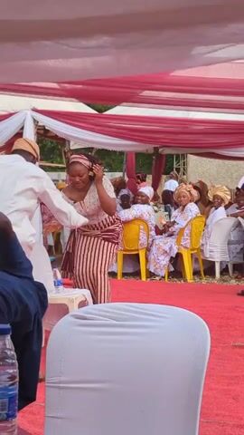 A woman in a cream-colored top and striped skirt dances on a red carpet under a white and red tent, facing the camera and then turning to her right. Another man in a white tunic and pants dances behind her to the left. Other people are seated at tables in the background, watching the performance. Some plastic water bottles and a white chair are in the foreground, out of focus.