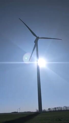 The video shows a wind turbine from a low angle against a bright blue sky. The sun is directly behind the turbine, creating a strong solar flare effect. The wind turbine has three blades that are turning slowly. The turbine is situated in a green field, and there are other turbines visible in the distance. There is a line of trees on the horizon.