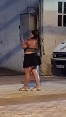 A woman with long, dark hair is standing on a paved surface at night. She is wearing a patterned off-the-shoulder top and a black skirt. She is holding a small object in her right hand and appears to be looking at it. Her left hand is also raised. Her body is angled to the right. The background is a building with a window and a utility box. To the right, a portion of a white vehicle is visible. Another woman, also with long dark hair, walks into the frame from the left, partially obscuring the first woman. She is wearing a patterned skirt and dark shoes. The lighting suggests an outdoor urban environment at night.