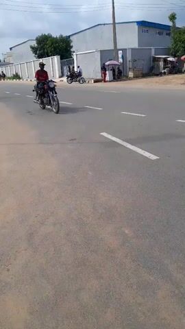 The video captures a scene on a paved road in Dam Camp, Nigeria. A person is riding a motorcycle in the foreground, traveling away from the camera. The motorcycle is a typical street bike, and the rider is wearing a red shirt and a dark cap. In the background, a long, light-colored building with a blue roof suggests an industrial or commercial area. A white wall with a fence runs alongside the road, separating it from the building. Further in the background, there are a few small stalls or shops, one with a pink umbrella, indicating a commercial area with street vendors. Some people are visible near these stalls. The sky is overcast, suggesting a cloudy day, possibly in the morning or afternoon given the shadows. The overall atmosphere appears to be that of a typical day in this location, with a mix of transport and commercial activity. The road has white lane markings, and the pavement shows some signs of wear. The camera is positioned from a low angle, looking slightly upwards. The video is a short clip, with no discernible sound.