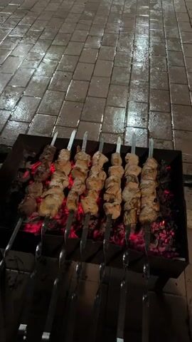 The video begins with a close-up of several skewers of meat cooking over glowing coals in a charcoal grill. The camera then pans up to reveal a paved area with three cars parked on it: a black sedan, a dark SUV, and a white SUV. The pavement is wet, reflecting the lights from overhead. In the background, under a trellis structure, a group of men are standing and talking. The scene suggests an outdoor gathering, possibly a barbecue, at night.