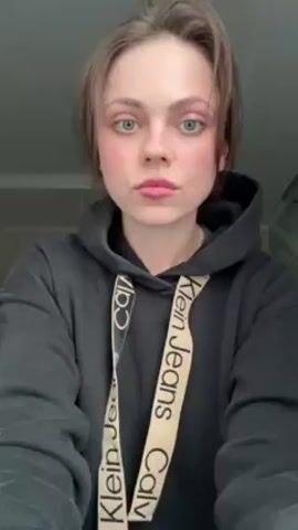 A young woman with light skin, light eyes, and light brown hair is centered in the frame, wearing a dark gray Calvin Klein hooded sweatshirt. She puckers her lips slightly, appearing to make a kissing face towards the camera. The background is nondescript, with what appears to be a slightly mottled wall behind her.