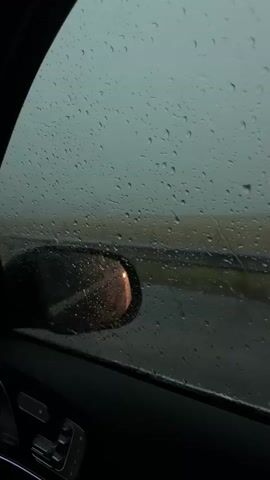 The video shows a close-up view through a car window during a rainstorm. Raindrops are visible on the glass, distorting the view of the outside. A side-view mirror is prominent on the left side of the frame, reflecting a sunset or sunrise. Beyond the mirror, a blurred landscape with a road and a signpost can be seen. The interior of the car, including part of the door panel and window controls, is visible in the foreground. The lighting is dim and overcast, consistent with rainy weather.