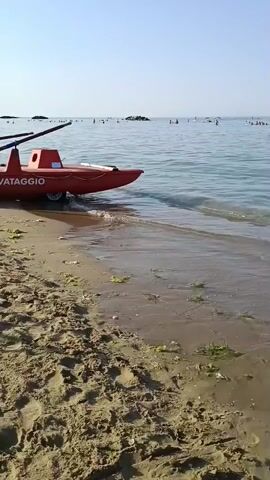 This is a video clip of a beach scene, likely filmed in Roseto degli Abruzzi, Italy, given the textual cues. The shot is taken from the sandy shore, looking out towards the ocean under a clear, bright blue sky, suggesting a sunny day.

The primary subjects are the sea and the beach. The water is calm with gentle waves lapping the shore. Scattered throughout the water, and closer to the beach, are several people swimming and enjoying the water. Some are further out, while others are closer to the shore, engaged in various water activities. There are also some inflatable objects visible in the water, such as a pink and blue ring.

In the foreground, on the wet sand where the waves have receded, there are small pieces of seaweed scattered. On the drier sand, further up the beach, there are some beach umbrellas visible in the distance, and a red rescue boat is partially visible on the left side of the frame. The boat has white wheels, suggesting it can be launched easily. Text on the boat reads "SALVATAGGIO," which translates to "RESCUE" in Italian, confirming its purpose.

Further out in the sea, there are some man-made structures, likely breakwaters, that extend from the sea. In the middle ground, a white signpost stands in the water. The overall atmosphere appears to be one of a pleasant summer day at the beach, with people relaxing and enjoying the seaside. The time of day appears to be morning or early afternoon, given the bright sunlight and clear sky.