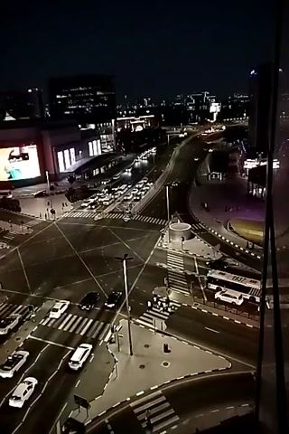 The video shows an aerial view of a busy intersection at night. Cars are driving through the intersection, and there are pedestrian crossings. A large building with a bright red advertisement is visible on the left side of the screen. The sky is dark, indicating it is nighttime. The intersection appears to be in a city, with lights from buildings in the background.