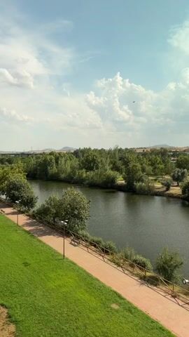 This video shows a panoramic view of a river and its surroundings. The river is wide and calm, with a slight ripple on its surface. On the left side of the river, there is a green grassy area and a dirt path lined with a wooden fence and lampposts. Lush green trees and bushes line the riverbank. In the distance, a town with buildings and trees can be seen. A stone bridge with several arches crosses the river in the far distance. The sky is blue with scattered white clouds. The overall scene is peaceful and scenic, depicting a natural landscape with man-made elements.