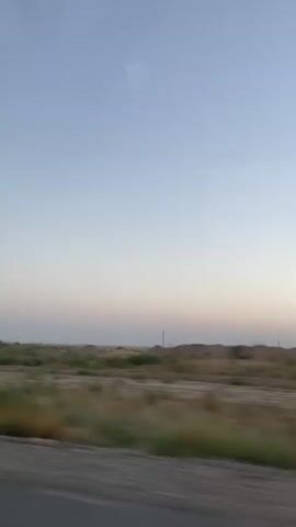 The video is a blurry, motion-filled shot from a vehicle traveling through a dry, grassy landscape under a pale blue sky. In the distance, there are a few scattered structures, possibly small buildings or utility poles. The foreground is dominated by the blurred impression of a road or path. The overall impression is one of vastness and a somewhat desolate environment.