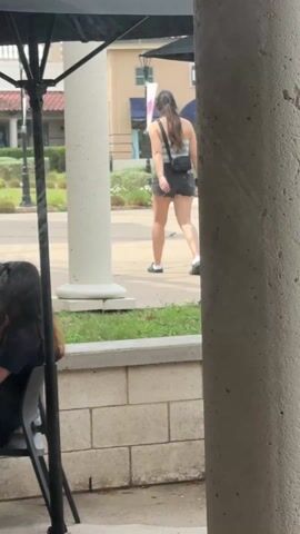 A woman walks away from the camera on a sidewalk with her dog on a leash. She wears a light-colored top, dark shorts, and dark shoes with white socks. She also has a small black bag over her shoulder. The dog appears to be short with light brown fur. The scene is outside, possibly a shopping area, as indicated by the storefronts, planters, light fixtures, and white concrete columns. A person sits at a table under an umbrella on a patio in the foreground. A second column is visible on the right.