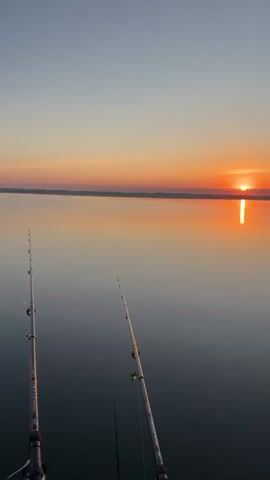 The video shows a sunrise over a calm body of water. Two fishing rods are visible in the foreground, positioned at an angle to capture the sunrise. The sun is a bright orange orb just above the horizon, casting a shimmering reflection on the water. The sky transitions from a light blue at the top to shades of orange and pink near the horizon. The water is still and reflects the colors of the sky, creating a mirror-like effect. The overall scene is peaceful and serene, suggesting an early morning fishing excursion.