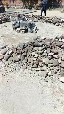 A person stands in an outdoor archaeological site, holding a phone to their ear and mouth, appearing to be on a call. The site is partially excavated, with low walls made of stones and bricks visible in a circular arrangement. Piles of rocks and debris are scattered around the area. In the background, there are trees and a building. The ground is covered in dirt and gravel. The sky is clear and blue.