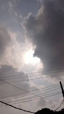 The video shows a view of the sky with white and dark gray clouds, through which the sun is visible. There are power lines in the foreground and a small patch of silhouetted foliage at the bottom of the frame.  Briefly, a blue, blurry, net-like object moves into view.  A house is visible in the final frame.