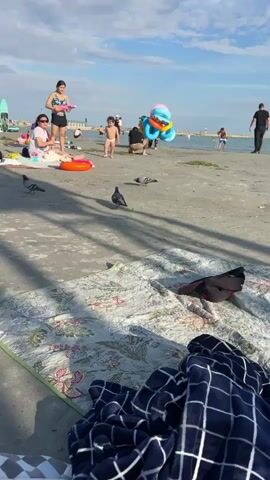 The video shows a beach scene with several people enjoying the day. In the foreground, a blanket with a floral pattern and a navy blue checkered blanket are laid out on the sand. Two pigeons are visible on the sand. In the mid-ground, there are several groups of people. One woman is holding a pink water gun, and a child is standing nearby. Another person, possibly a child, is playing with colorful balloons. Further in the background, there are more people, beach umbrellas, and surfboards. The sky is partly cloudy.