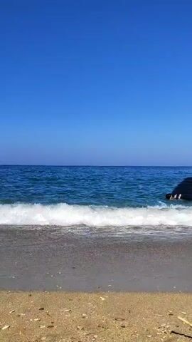 The video opens to a view of a sandy beach with the ocean stretching out under a clear blue sky. Small waves are breaking on the shore. In the distance, a person is in the water with a black inflatable object. The person moves into the water and appears to be holding onto the inflatable object. The video continues with the person and the object floating in the water as the waves continue to lap at the shore.