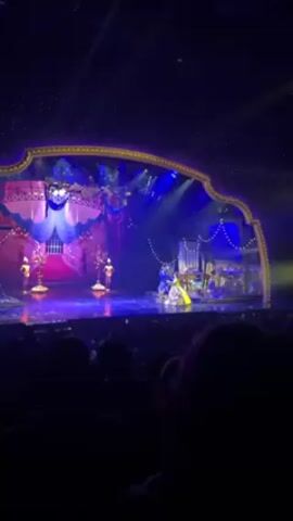 Here is a description of the video:

The video shows a scene from a theatrical performance, likely "Beauty and the Beast." The stage is decorated with a grand staircase, ornate props like candelabras, and shimmering backdrops.  A performer dressed as Belle in a gold and yellow gown is on stage. She begins to dance with a performer in a blue and gold costume, portraying the Beast. They move gracefully together across the stage. An audience can be seen in the foreground of the video. The scene is bathed in blue and purple lighting.