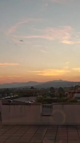 The video shows a panning view of a scenic landscape at sunset. The sky is filled with soft pink and orange clouds, with mountains visible in the distance. In the foreground, there is a building with a tiled rooftop and a balcony. There are also trees and some parked cars visible in the midground. The overall impression is one of a peaceful and tranquil evening.