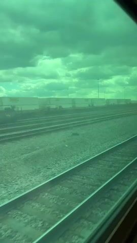 The video shows a view from a train moving alongside a railway yard. Several parallel tracks run alongside the train.  Freight cars, stacked shipping containers, and utility poles are visible in the background. The sky is overcast, and everything is cast in a green hue, possibly due to tinted glass in the train window.