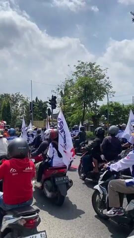 This video captures a daytime scene on a bustling street in Tebel, Indonesia, under a sky with scattered clouds. A large group of people are participating in what appears to be a protest or procession, primarily riding motorcycles.

The main subjects are numerous individuals, mostly men, mounted on motorcycles, wearing helmets. Several flags are prominently displayed, some white with a red and blue logo that includes the text "FSPMI," and others red with a white logo. In the center of the road, a truck fitted with large speakers and what looks like a makeshift platform is central to the procession, with people standing on it and seemingly addressing the crowd.

The motorcycles are stopped at a traffic light, which is currently yellow. The overall atmosphere suggests a united front, possibly heading towards a specific destination.

Notable details include the bright, sunny weather. Visible motorcycle license plates such as "W 6598 NDE," "W 5610 XD," and "W 2030 NCS" confirm the Indonesian setting, specifically the Surabaya/Sidoarjo region in East Java. A person in the foreground wears a red hoodie with "Fre ship" and "2017" printed on the back. Other riders wear shirts also bearing the "FSPMI" logo.

An audio snippet provides crucial context, with a voice saying: "Ayo jalan, cepat Jumat'an, jangan masuk, kantor kejaksaan." This translates to "Let's go, hurry for Friday prayer, don't enter, Prosecutor's Office," suggesting the group is engaged in a demonstration, potentially near or en route to a government or legal institution, with an instruction about an upcoming Friday prayer and a directive regarding the Prosecutor's Office.