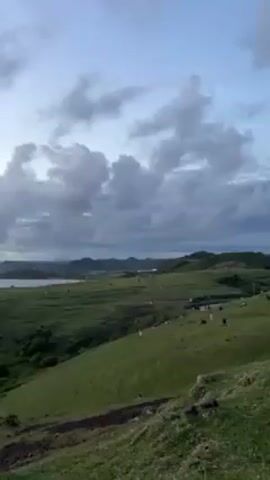 The video shows a panoramic view of a coastal landscape with a grassy foreground, a beach and ocean in the middle ground, and distant hills under a cloudy sky. The sky transitions from a dusky blue and grey to orange hues as the video progresses, indicating a sunset or sunrise. There are people visible on the grassy area and beach, though they appear small in relation to the landscape. The camera slowly pans from left to right, highlighting the various features of the scene.