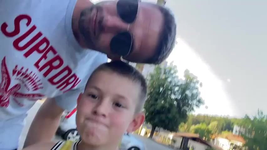 This is a short video that appears to be a selfie taken outdoors, likely in Gabrovo, Bulgaria. The scene features two people, an adult male and a young boy. The adult is wearing a light blue t-shirt with red lettering that reads "SUPERDRY". He has dark hair, a beard, and is wearing sunglasses. The young boy is in the foreground, wearing a yellow tank top with black lettering that says "NYC 82". He has short, dark hair and is looking at the camera with a big smile, extending his hands towards the lens as if playing or being playful. The background shows a street or pathway, some trees, and buildings, suggesting a residential or suburban area. There is a wooden pole visible behind the adult. The lighting suggests it is daytime, likely late afternoon or early evening, with bright, diffused sunlight. The overall mood seems to be lighthearted and playful.