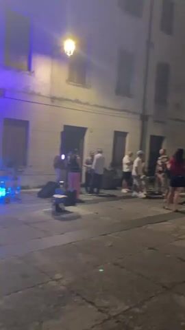 The video shows a nighttime street scene in a European town or city. A group of people are gathered on a cobblestone street, with buildings lining both sides. Some people are standing and talking, while others are sitting at tables outside what appears to be a cafe or restaurant. There is a street lamp illuminating the area, and a few speakers are visible on the ground. The atmosphere seems casual and social, with people enjoying themselves outdoors in the evening. The architecture of the buildings suggests an older, possibly historic, setting.