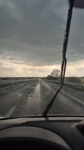 The video shows the perspective of someone driving on a highway during rainy weather. The windshield wipers are actively clearing the rain. The sky is overcast with dark clouds. The road is wet and stretches out into the distance. A few vehicles are visible in the distance and a tree is seen on the side of the road. The dashboard of the car is visible in the foreground.
