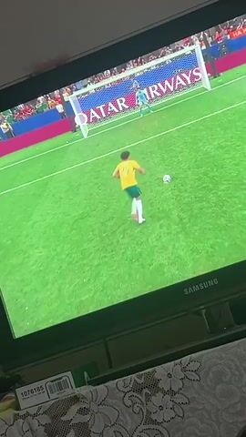 A young male soccer player with curly hair and a yellow jersey with the number 25 on the back, white shorts, and white socks is shown on a television screen. He is on a green soccer field with a soccer net in the background. The net has a banner that reads "QATAR AIRWAYS." The player appears to be dribbling or moving the ball, with a soccer ball visible on the field near him. The television screen displays the game in progress, and there is some text on the screen indicating the player's name is "HERRINGTON." The image is captured from an angle, likely from a phone recording the television.
