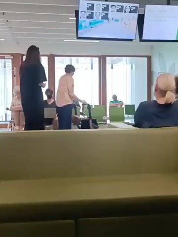 The video shows a waiting room. In the foreground, a green sofa takes up the bottom portion of the frame. In the middle ground, several people are seated on green chairs. A woman in a black dress stands facing away from the camera. To her right, a person in a tan shirt bends over a bag, and to their right, another person is seated. To the left of the woman in black, a person sits in a wheelchair and looks to the left, and another person sits with a laptop. In the background, there are large windows and doors. Above the windows, a large screen displays images of people and some text.