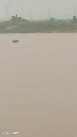 The video shows a wide, still river under an overcast sky. The water is a light, muddy brown color. On the far bank of the river, there's a dense line of green vegetation, possibly trees and shrubs. In the distance behind the vegetation, utility towers are visible, silhouetted against the gray sky. A small boat with one or more people in it can be seen on the water. Tall blades of grass are in the bottom right corner of the frame. The scene is still and quiet, suggesting a remote or rural location. An "Infinix NOTE 11i" watermark is visible at the bottom of the screen.