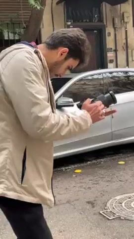 A young man with dark hair and a beard is standing on a sidewalk, holding a digital camera in his hands. He is wearing a light beige jacket, a red and black plaid shirt, and dark pants. He appears to be reviewing photos or videos on the camera's screen. In his right hand, he holds a lit cigarette. Behind him is a silver Mercedes-Benz parked on the street. A manhole cover is visible on the sidewalk near his feet. There are trees and a building in the background. The lighting suggests it is daytime, possibly overcast.