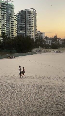 A medium shot shows a couple walking across a sandy beach against a backdrop of tall, modern buildings under a dusky sky.  The couple, a male and a female, are walking at a leisurely pace; the male is wearing a white shirt, dark shorts, and a cap, and he is carrying a white object. The female is wearing a dark dress. The sandy beach is predominantly white, with visible footprints. Behind the beach, there's a low fence and some trees. Further back are several multi-story buildings, casting silhouettes against the slightly orange and pink tinted sky. A few individuals are seated together at the end of the frame behind the fence.