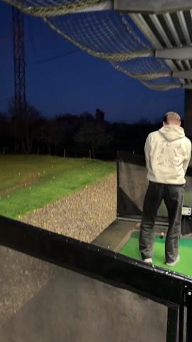 At dusk, a person in a light-colored hoodie and dark pants is seen at a golf driving range. The person stands on a green mat within a booth, holding a golf club. The booth has a black frame and is partially enclosed by a wire net overhead. Beyond the booth, a grassy field is visible, dotted with yellow golf balls, extending towards a treeline against a dark blue sky. A tall metal tower stands in the distance. The person takes a swing and hits a golf ball. The camera briefly follows the ball's trajectory before returning to the view of the field.