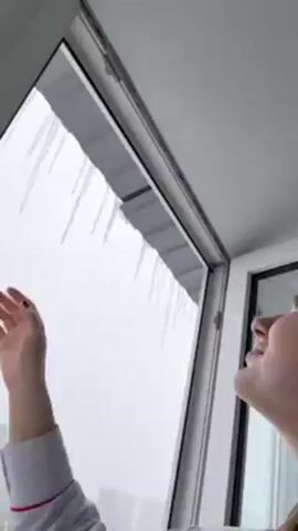 A woman in a gray and white striped pajama top breaks off an icicle from a row of icicles hanging off an outside overhang, visible through a white-framed glass window. She looks at the camera and then back up to break off another icicle. The environment suggests it is winter, with a snow-covered landscape outside the window. Her hair is pulled back with a white cloth, and she has light makeup on.