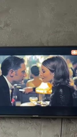 A close-up shot shows two people, a man and a woman, facing each other with their lips touching in a kiss. They are seated at a table in what appears to be a dimly lit restaurant or ballroom, with other patrons blurred in the background. A champagne glass is visible on the table between them. The man is wearing a dark suit and tie, and the woman is wearing a dark, sparkly top. The scene suggests an intimate moment between the couple.