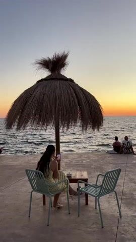 A woman sits at a small table on a paved area near a body of water, looking at her phone. Two chairs face the table, one occupied by the woman, the other empty. A large thatched umbrella stands behind the table, providing shade. In the background, the ocean meets the horizon under a sky transitioning from dusk to night. Further in the distance, two people are seated on the ground, facing away from the camera. The overall scene depicts a tranquil evening at a beachside location.