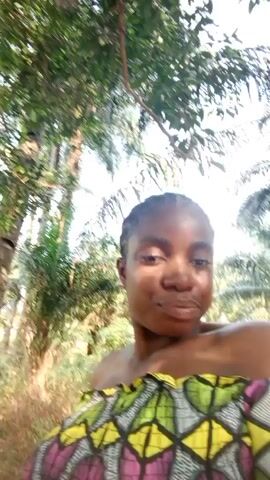 The video shows a person standing outside in an area with trees and dirt. The person has brown skin, dark eyes, and dark hair pulled back. They are wearing a colorful off-the-shoulder top. The camera angle is low, looking up at the person's face. The background is slightly blurred, showing trees and foliage. As the video progresses, the camera moves, and the person walks into a clearing where there is more dirt and some debris. The person maintains eye contact with the camera throughout the video.