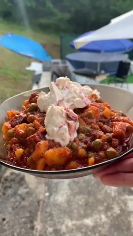 The media file shows a close-up of a bowl of food, held by a person's hand. The food appears to be a stew or casserole, with chunks of potatoes, peas, and a reddish sauce, topped with dollops of a white creamy substance, possibly sour cream or yogurt, with some red flecks. The background suggests an outdoor setting, possibly a picnic or camping area. There are glimpses of trees and greenery, and in the distance, parts of tents or umbrellas, one white and blue. The lighting indicates it is daytime, likely late afternoon or early evening, with the sun casting rays. The overall mood seems relaxed and casual. The location is identified as Novo Bardo, Bulgaria. There is no visible text in the media file.