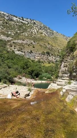 The video shows a natural setting with a rocky waterfall and a pool of water. Several people are present, some are bathing in the pool while others are relaxing on the rocks. One person in the foreground is kneeling on the rocks, appearing to be taking a photograph or video. The surrounding environment is characterized by lush green vegetation, rocky cliffs, and a clear blue sky.