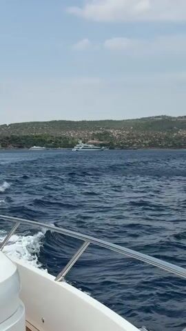 This video shows a view from the side of a boat moving through choppy, dark blue water. In the distance, a hilly, green coastline is visible under a cloudy sky. Several large white yachts are anchored or moving slowly in the water off the coast. The edge of the boat's railing and white hull are in the foreground.
