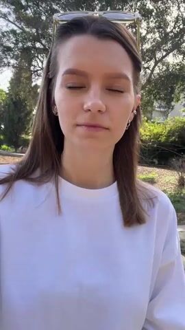 A young woman stands in a park on a sunny day near a beach. She has shoulder-length brown hair, is wearing a white t-shirt, and has sunglasses on top of her head. She smiles at the camera and looks around at the scenery. The park has trees, bushes, and a paved pathway. In the background, the beach is visible with people enjoying the water. The sky is blue, and the sun is shining brightly.