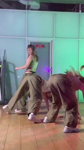 Three women are dancing together indoors. They are wearing crop tops and baggy, olive-green pants. Their hair is long and loose. Two women are bent forward at the waist, supported by their hands, while the third is upright and dancing while facing the camera. The background is a glossy wooden floor and a paneled wall with a door. There is green lighting coming from the right. There is a metal pole in the background on the left.