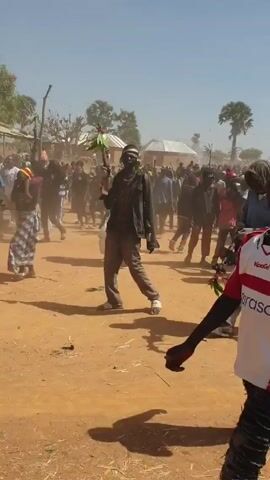 The video shows a crowd of people on a dirt road in a rural setting. The environment appears dry and dusty. Many of the people are moving around, some walking, some dancing. A few people are dressed in what appear to be costumes, one with a mask. There are explosions that generate white smoke. There are houses visible in the background, and some trees. Several children are visible in the foreground, including one wearing a red and white soccer jersey. Overall, the scene appears chaotic, with a sense of celebration or unrest.