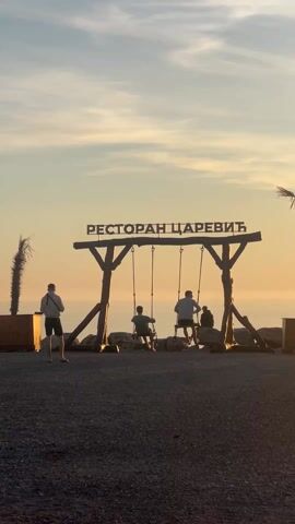The video shows a tranquil scene at sunset, featuring the "Restaurant Carevic" sign displayed above a set of swings. Two people are sitting on the swings, enjoying the view, while a third person stands to the side, observing. The background showcases a beautiful sky with soft, warm hues and the sea stretching out towards the horizon. The area around the swings is covered with gravel.