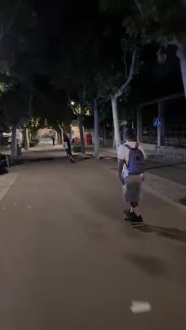 A person on a skateboard is filmed from behind at night on a paved path. The person is wearing a blue hat, a white t-shirt, and patterned shorts, with a purple backpack. Another person is visible further down the path, also on a skateboard. The path is lined with trees and streetlights. The scene appears to be an urban park or promenade.