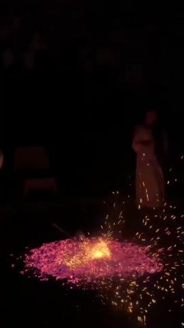 A dark outdoor scene at night features a pile of glowing red embers on the ground. Sparks are emanating from the embers. A person in a light-colored robe stands to the right of the embers, facing them. To the left of the embers, another person in a light-colored shirt and dark pants is seen with their back to the camera, holding a long, thin object that appears to be interacting with the embers, possibly stirring them. In the background, the silhouette of trees and foliage is visible against the dark sky, and to the far left, the shape of what might be a chair or bench is faintly discernible. The overall impression is of a ritualistic or celebratory event involving fire.