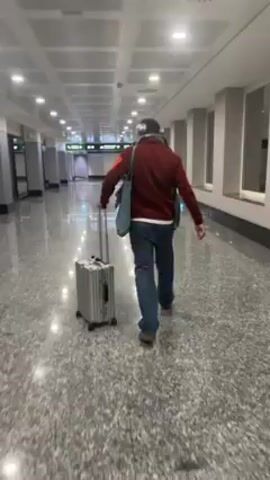 The video shows the interior of an airport terminal hallway with pale marble floor. A man is seen from the back pulling a small, silver suitcase. He is wearing a maroon colored coat, jeans, and a light colored hat. He also has a pale blue tote bag over his shoulder. Another man in a dark blue hoodie walks by. The terminal appears to be mostly empty with the occasional passerby. There are green directional signs overhead and large white pillars along the sides of the hallway.