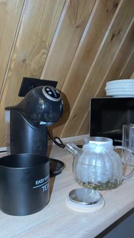 The video shows a countertop in a room with wood-paneled walls. On the countertop are a black coffee machine, a black cup with "EASY MORNING TEA" printed on it, a glass teapot with dried tea leaves inside, and a small silver and wood lid. A microwave is visible in the background. Water is being poured from an unseen source into the glass teapot.